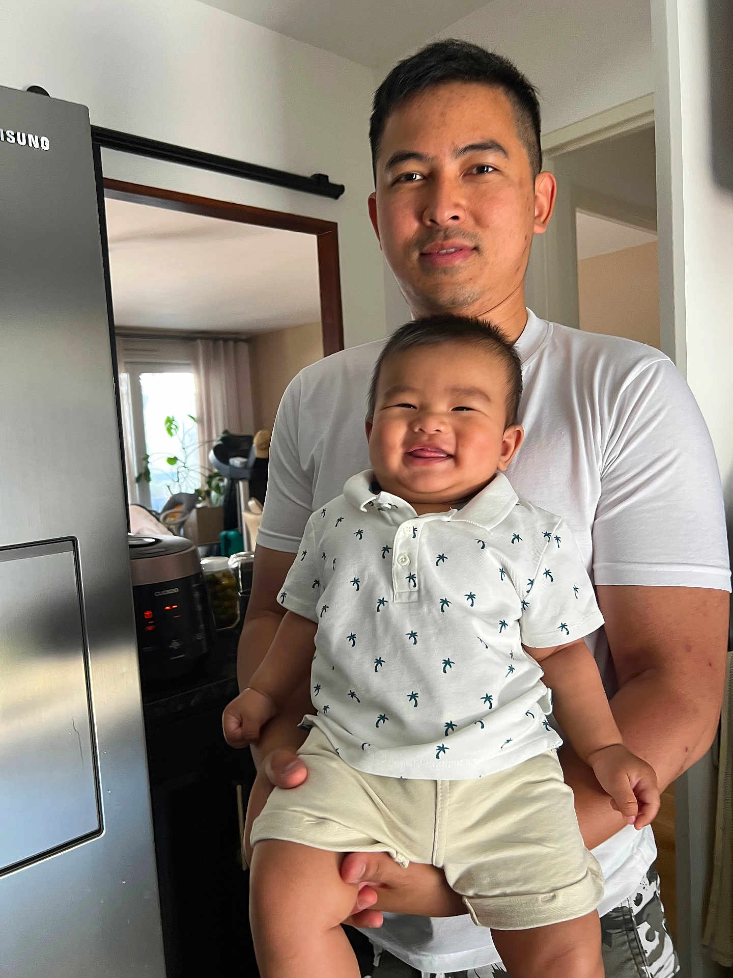 Sak a rejoint le concours — aidez-le/la à gagner de superbes lots ! baby, adult, smile, person, indoor, kitchen, clothing, holding, shorts, shirt, refrigerator, appliance, cabinet, plant, window, curtain, happy, family, portrait, casual