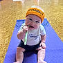 Roan is registered to the contest to win money with this photo: baby, child, headband, orange, purple_yoga_mat, yoga, indoor, floor, people, sitting, chewing, clothing, shorts, tshirt, person, infant, exercise, group, mat, playful