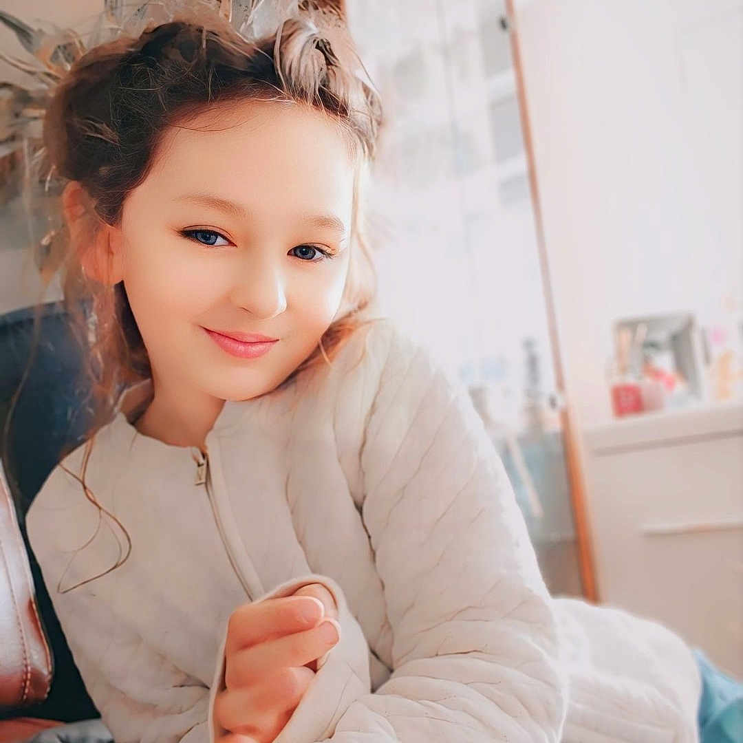 Inaya a rejoint le concours — aidez-le/la à gagner de superbes lots ! candid, casual_clothing, child, childhood, comfort, cozy, face, furniture, home_interior, indoor, long_hair, person, plant, portrait, quilted_surface, relaxed, smile, soft_lighting, white_jacket, young_person