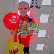 Kenzo participe au concours pour gagner de l'argent avec cette photo : babybel, bag, black_pants, boy, celebration, certificate, child, diploma, door, elf_decoration, floor_mat, frisbee, happy, indoor, m&m, red_jacket, smiling, sneakers, standing, toy