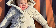 Amelia joined the competition — help win amazing prizes! baby, child, snowsuit, winter_clothing, hood, mittens, smile, happy, sitting, outdoor, cold_weather, cute, infant, padded_jacket, rosy_cheeks, blue_eyes, playful, striped_background, warm_clothing, portrait