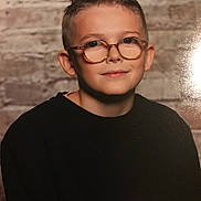 Rayan participe au concours pour gagner de l'argent avec cette photo : child, boy, glasses, portrait, smile, short_hair, black_shirt, brick_wall, indoor, casual, face, head, young, eyewear, fashion, person, human, background, warm_lighting, studio_photo