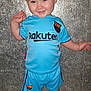 child, toddler, smiling, blue_clothing, soccer_kit, flip_flops, standing, indoors, floor_tiles, shorts, tshirt, person, cute, happy, face, hair, skin, footwear, sport, background_wall
