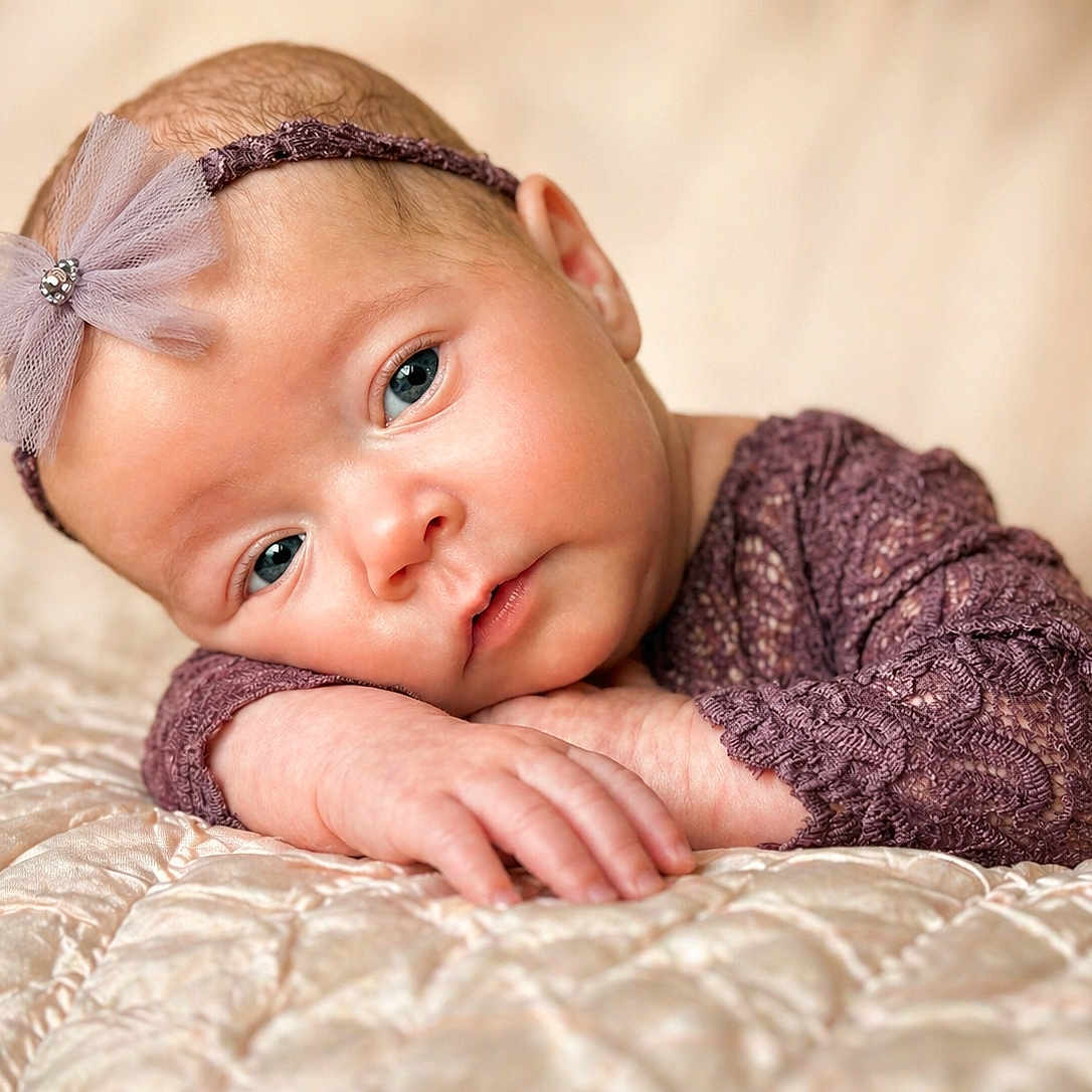 Meilani is registered to the contest to win money with this photo: baby, infant, face, headband, bow, purple_clothing, lace, blanket, quilted, cute, portrait, lying_down, skin, small_hands, blue_eyes, soft_light, close_up, indoors, resting, newborn