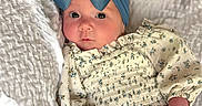 Meilani is registered to the contest to win money with this photo: baby, blanket, blue_bow, child, cute, expression, face, floral_dress, hands, headwear, indoor, infant, lying_down, person, portrait, skin, soft_light, texture, white_leggings, young