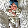 baby, blue_bow, child, curious_expression, cute, face, floral_pattern, headwear, indoor, infant, long_sleeves, onesie, person, portrait, sitting, small_hands, soft_lighting, textured_blanket, tights, white_blanket