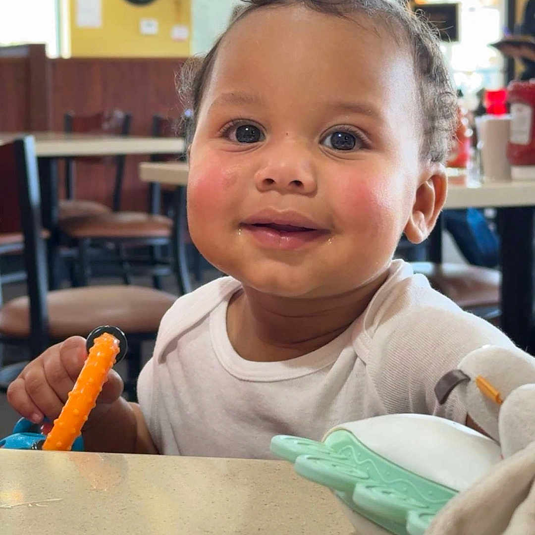 Darann is registered to the contest to win money with this photo: background, blush, chairs, child, condiments, curly_hair, face, furniture, happy, indoor, light, person, restaurant, smiling, table, teething_toy, toddler, toy, white_shirt, window