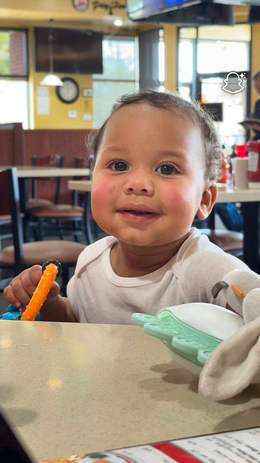 Darann is registered to the contest to win money with this photo: toddler, child, smiling, indoor, restaurant, table, toy, face, person, happy, curly_hair, white_shirt, furniture, chairs, condiments, blush, teething_toy, background, window, light