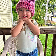 Lilly is registered to the contest to win money with this photo: child, girl, smiling, hat, knitted_hat, pink, shorts, denim, butterfly_print, shirt, outdoor, porch, chair, wood, greenery, grass, happy, person, cute, summer