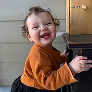 Nemi is registered to the contest to win money with this photo: child, toddler, smiling, curly_hair, orange_sweater, black_skirt, hand, wooden_table, indoor, face, happy, cute, person, portrait, baby, clothing, furniture, skin, background, expression