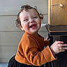 child, toddler, smiling, curly_hair, orange_sweater, black_skirt, hand, wooden_table, indoor, face, happy, cute, person, portrait, baby, clothing, furniture, skin, background, expression