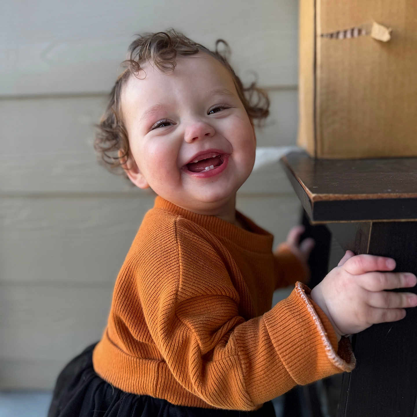 Nemi is registered to the contest to win money with this photo: child, toddler, smiling, curly_hair, orange_sweater, black_skirt, hand, wooden_table, indoor, face, happy, cute, person, portrait, baby, clothing, furniture, skin, background, expression