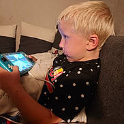 Ryan participe au concours pour gagner de l'argent avec cette photo : child, blonde_hair, gaming_device, couch, indoor, focused, black_shirt, polka_dots, blanket, playing, game_console, young_boy, technology, relaxed, seated, casual, home_interior, side_profile, entertainment, cord