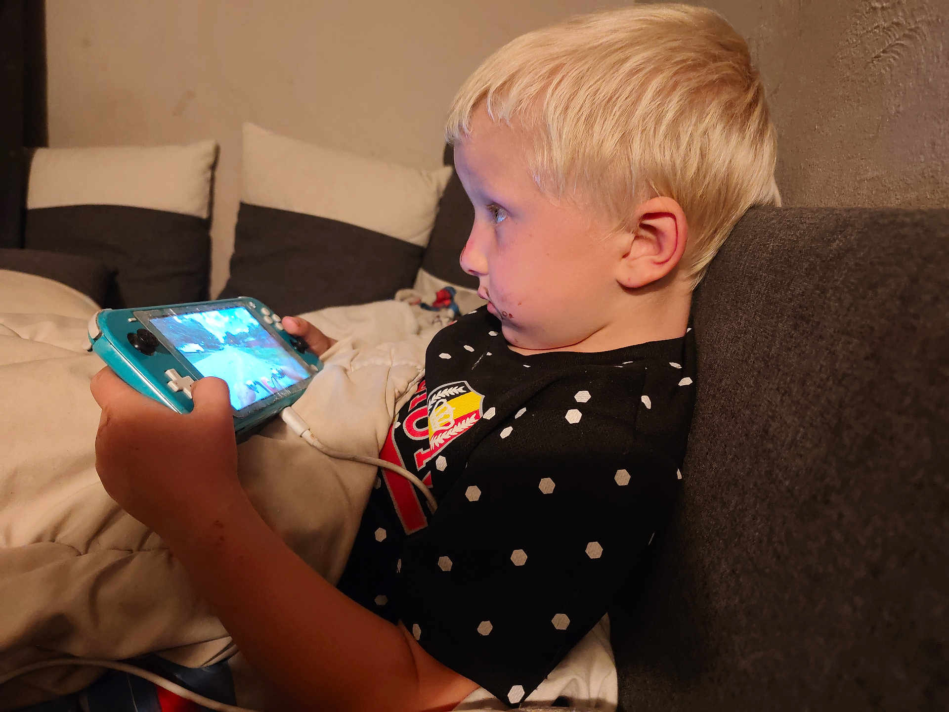 Ryan participe au concours pour gagner de l'argent avec cette photo : child, blonde_hair, gaming_device, couch, indoor, focused, black_shirt, polka_dots, blanket, playing, game_console, young_boy, technology, relaxed, seated, casual, home_interior, side_profile, entertainment, cord