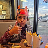 Angel is registered to the contest to win money with this photo: child, boy, portrait, smiling, thumbs_up, fast_food, french_fries, restaurant, burger_king, crown, paper_crown, table, seated, indoors, window, cars, evening, casual_clothing, happy, food