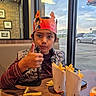 child, boy, portrait, smiling, thumbs_up, fast_food, french_fries, restaurant, burger_king, crown, paper_crown, table, seated, indoors, window, cars, evening, casual_clothing, happy, food