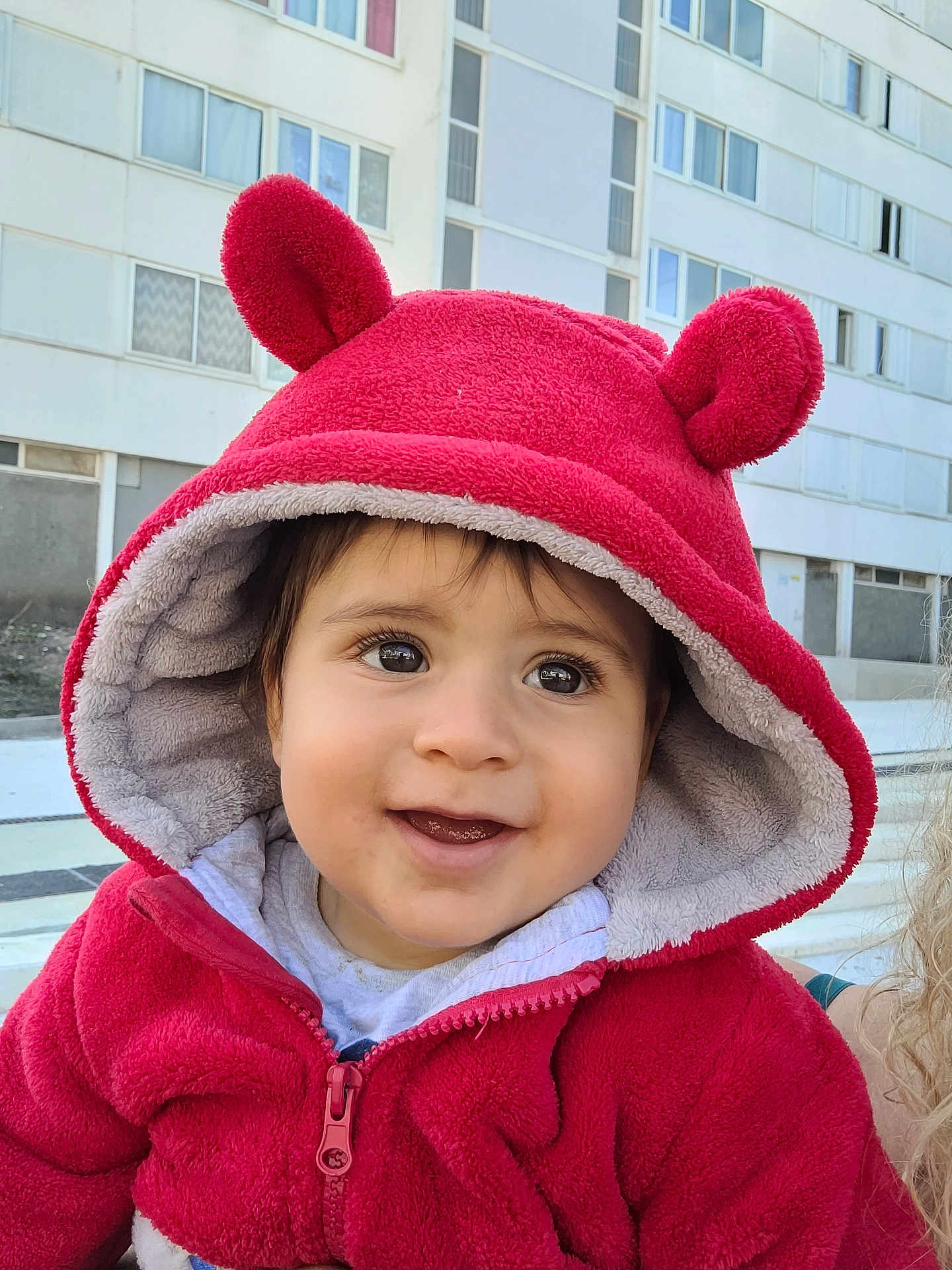 Kaël participe au concours pour gagner de l'argent avec cette photo : baby, child, hoodie, red_clothing, smile, face, outdoor, urban, building, window, zipper, cute, hood, eyes, person, portrait, warm_clothing, daylight, expression, young_child