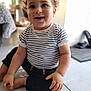 Kaël a rejoint le concours — aidez-le/la à gagner de superbes lots ! toddler, child, smiling, indoor, floor, striped_shirt, black_pants, curly_hair, happy, sitting, barefoot, person, portrait, cute, young_child, bright, natural_light, closeup, baby, casual_clothing