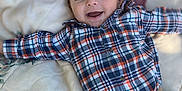 Jaxon is registered to the contest to win money with this photo: baby, child, smiling, plaid_shirt, pants, blanket, floral_pattern, lying_down, happy, portrait, indoor, cute, infant, person, clothing, baby_face, young_child, casual_wear, relaxed, soft_lighting