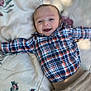 baby, child, smiling, plaid_shirt, pants, blanket, floral_pattern, lying_down, happy, portrait, indoor, cute, infant, person, clothing, baby_face, young_child, casual_wear, relaxed, soft_lighting