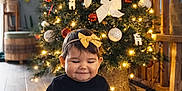 Soline a rejoint le concours — aidez-le/la à gagner de superbes lots ! baby, child, christmas_tree, holiday, decorations, ornaments, lights, bow, headband, black_clothing, floor, wooden_floor, indoor, festive, smiling, cute, holiday_season, toddler, seated, happy