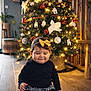 baby, child, christmas_tree, holiday, decorations, ornaments, lights, bow, headband, black_clothing, floor, wooden_floor, indoor, festive, smiling, cute, holiday_season, toddler, seated, happy