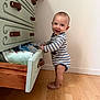 baby, child, striped_onesie, standing, barefoot, drawer, towels, wooden_floor, smile, happy, interior, furniture, cabinet, face, person, home, cute, infant, looking_at_camera, room
