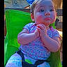 baby, background_objects, casual, child, clothing, colorful, cute, expression, face, furniture, green_chair, hands_clasped, indoor, patterned_pants, photography, portrait, seatbelt, short_sleeve_shirt, toddler, wooden_table