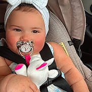Valentina is registered to the contest to win money with this photo: baby, child, car_seat, pacifier, headband, bow, plush_toy, stuffed_animal, clothing, dress, seatbelt, infant, cute, pink, white, hand, face, indoors, car, safety