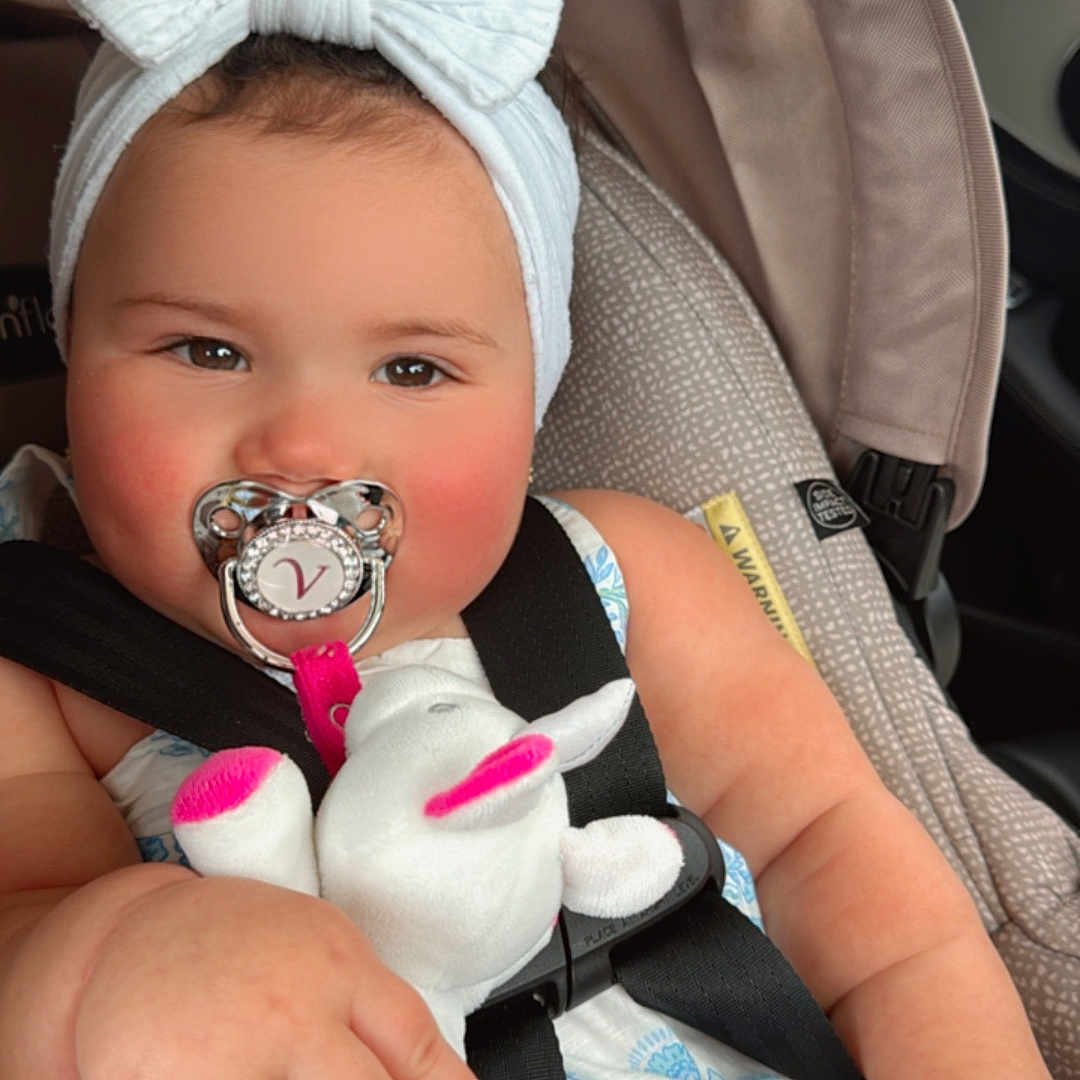 Valentina is registered to the contest to win money with this photo: baby, bow, car, car_seat, child, clothing, cute, dress, face, hand, headband, indoors, infant, pacifier, pink, plush_toy, safety, seatbelt, stuffed_animal, white