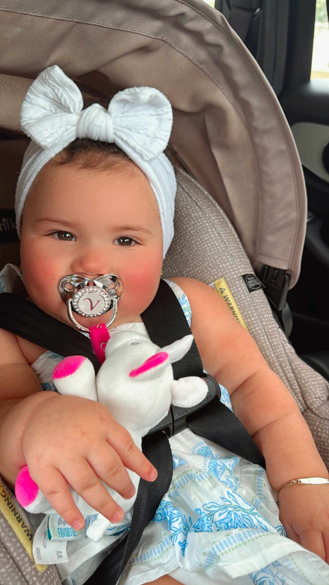 Valentina is registered to the contest to win money with this photo: baby, child, car_seat, pacifier, headband, bow, plush_toy, stuffed_animal, clothing, dress, seatbelt, infant, cute, pink, white, hand, face, indoors, car, safety
