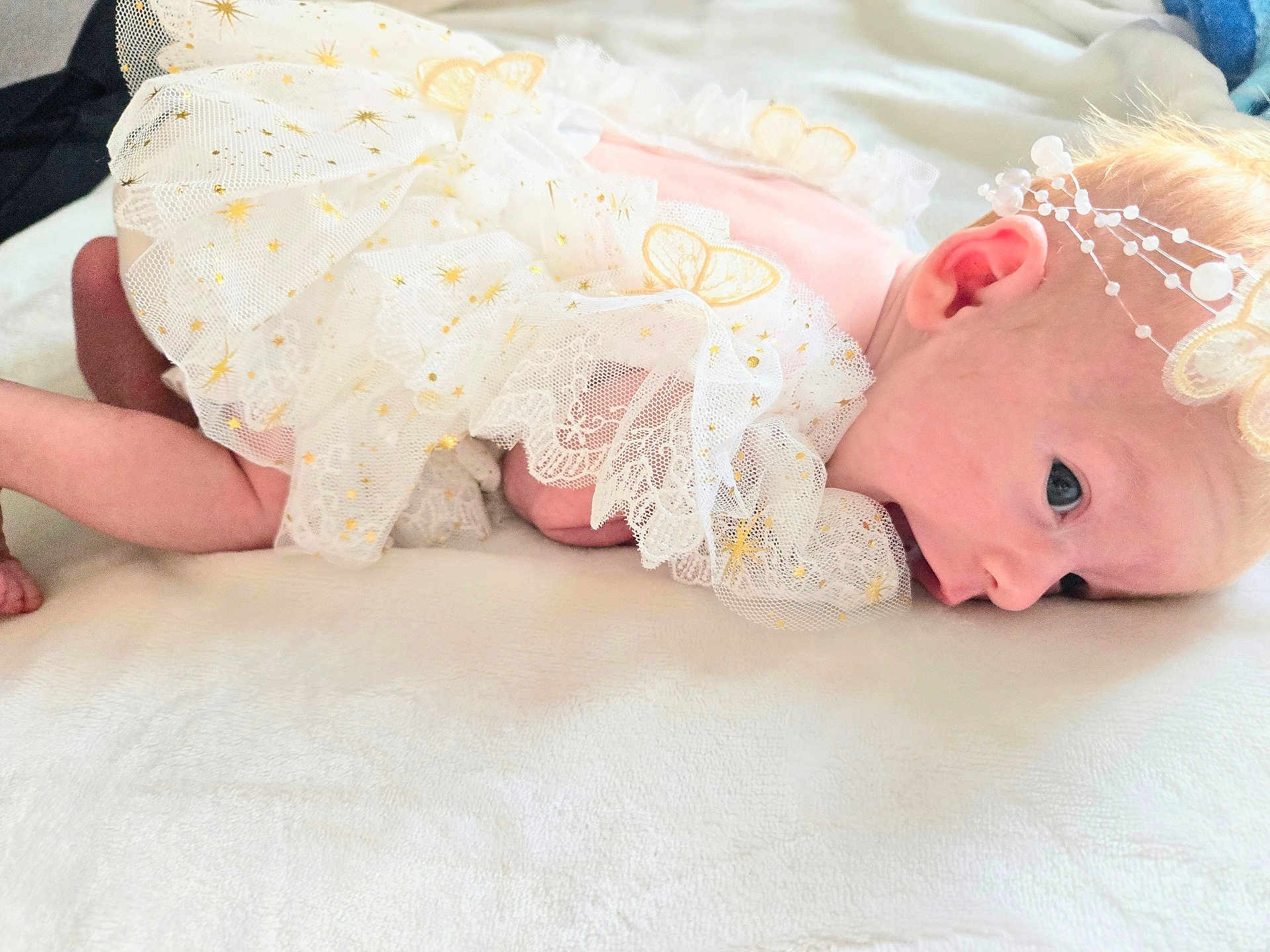 Eliana is registered to the contest to win money with this photo: baby, infant, frilly_dress, white_clothing, headband, pearl_accessory, lying_down, soft_surface, curious_eyes, newborn, cute, delicate, closeup, face, skin, small_hands, small_feet, indoors, blanket, portrait