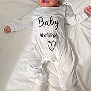 Adam participe au concours pour gagner de l'argent avec cette photo : baby, infant, smiling, white_onesie, lying_down, blanket, face, happy, child, indoors, cute, sock, portrait, newborn, soft_texture, text_on_clothing, arabic_text, bed, cozy, person
