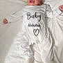 baby, infant, smiling, white_onesie, lying_down, blanket, face, happy, child, indoors, cute, sock, portrait, newborn, soft_texture, text_on_clothing, arabic_text, bed, cozy, person