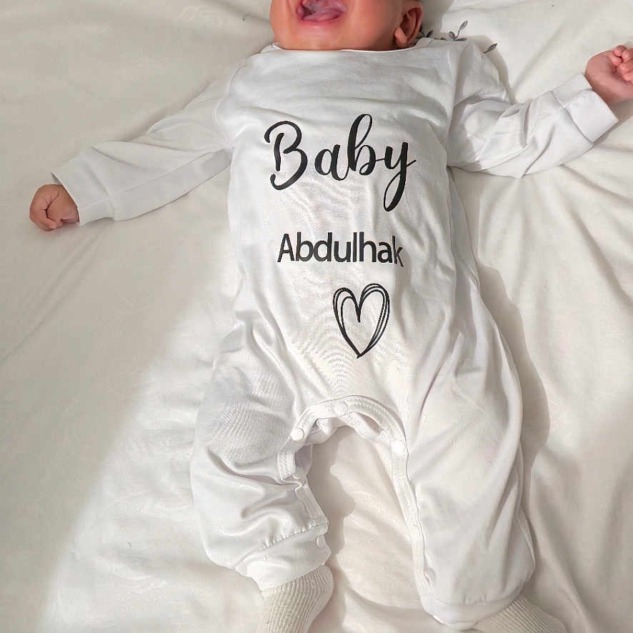 Adam participe au concours pour gagner de l'argent avec cette photo : arabic_text, baby, bed, blanket, child, cozy, cute, face, happy, indoors, infant, lying_down, newborn, person, portrait, smiling, sock, soft_texture, text_on_clothing, white_onesie
