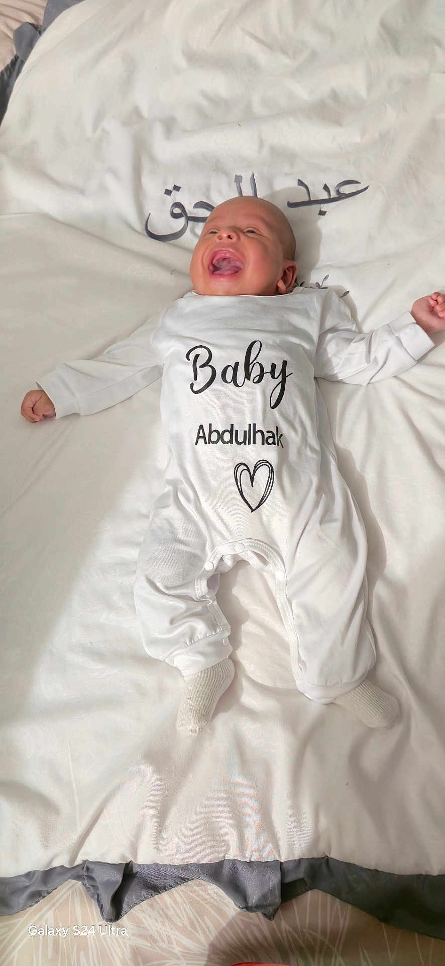 Adam participe au concours pour gagner de l'argent avec cette photo : baby, infant, smiling, white_onesie, lying_down, blanket, face, happy, child, indoors, cute, sock, portrait, newborn, soft_texture, text_on_clothing, arabic_text, bed, cozy, person