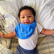 Amari is registered to the contest to win money with this photo: baby, smiling, blue_bib, curly_hair, white_pillow, bed, blanket, plaid_blanket, infant, happy, child, lying_down, cute, indoor, portrait, clothing, person, young_child, head, face
