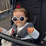 Kamélya participe au concours pour gagner de l'argent avec cette photo : toddler, child, sunglasses, pacifier, stroller, bow, hair, hand, clothing, white_shirt, baby_accessory, outdoor, seatbelt, black_stroller, cute, fashion, person, baby, walking, playful