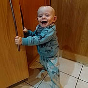 Kai is registered to the contest to win money with this photo: baby, child, kitchen, cabinet, wood, floor, tile, pajamas, smiling, happy, standing, holding, handle, clothing, indoor, young_child, foot, face, person, cute