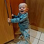 baby, child, kitchen, cabinet, wood, floor, tile, pajamas, smiling, happy, standing, holding, handle, clothing, indoor, young_child, foot, face, person, cute