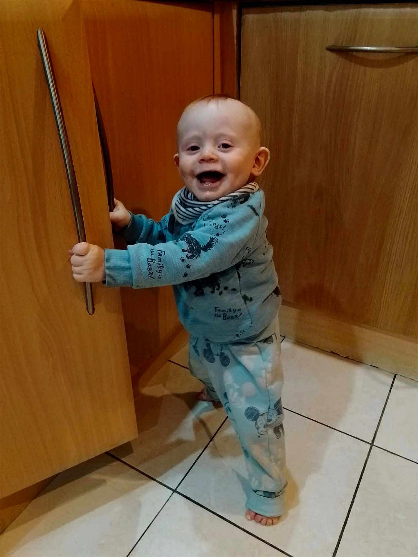 Kai is registered to the contest to win money with this photo: baby, child, kitchen, cabinet, wood, floor, tile, pajamas, smiling, happy, standing, holding, handle, clothing, indoor, young_child, foot, face, person, cute