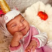 Sayda is registered to the contest to win money with this photo: baby, sleeping, unicorn_costume, costume, horn, pink_clothing, fluffy_blanket, pumpkin, orange, cute, infant, hand, soft_texture, cozy, portrait, indoors, child, smiling, closed_eyes, cute_outfit