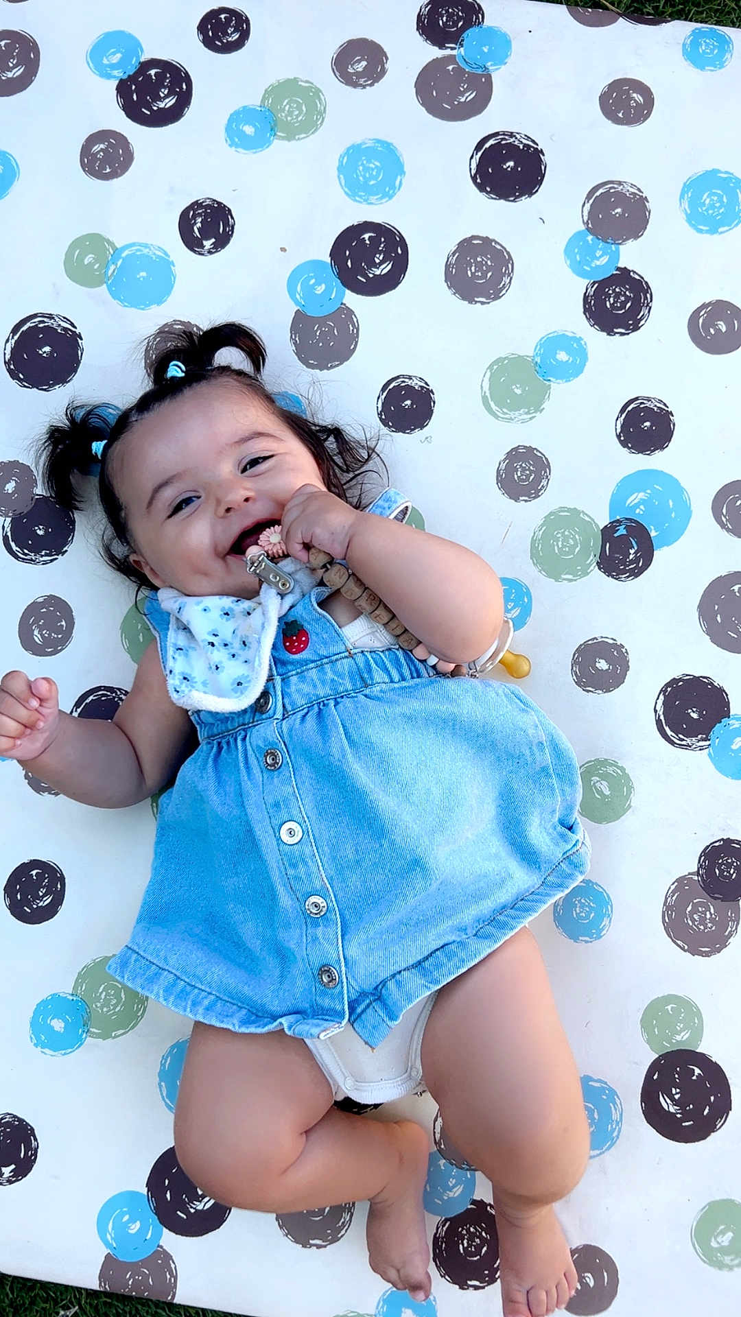 Andreïa participe au concours pour gagner de l'argent avec cette photo : baby, child, infant, smiling, happy, pigtails, blue_dress, polka_dot, toy, beads, playmat, outdoor, grass, cute, chewing, barefoot, lying_down, accessory, white_bib, buttoned_dress