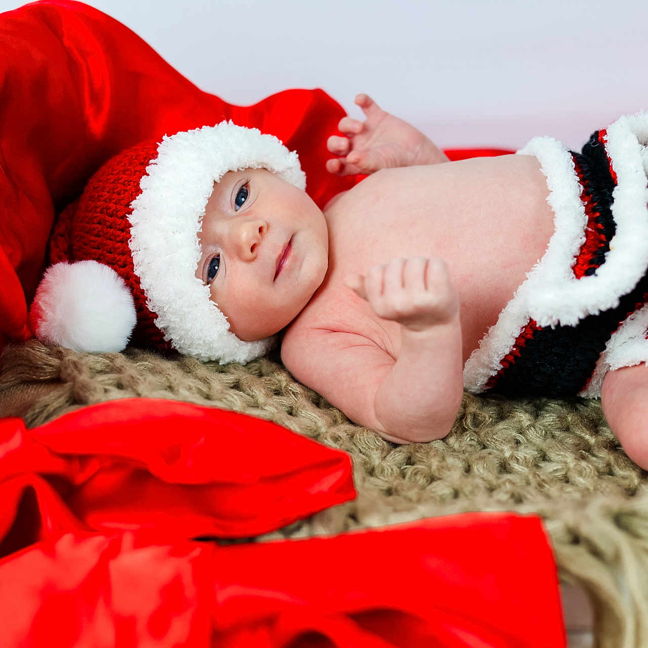 Kagen is registered to the contest to win money with this photo: baby, blanket, child, christmas, cozy, cute, face, festive, holiday, indoors, infant, knitted_clothing, lying_down, newborn, portrait, red_fabric, santa_hat, smiling, soft_texture, warm_colors
