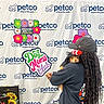 person, dog, costume, hat, dreadlocks, petco, sign, flowers, banner, event, celebration, cute, small_dog, black_clothing, backdrop, holding, pet, mother's_day, colorful, indoor