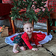 Serenity is registered to the contest to win money with this photo: baby, child, crawling, christmas_tree, holiday, festive, red_clothing, green_skirt, smiling, indoor, decorations, plaid, wooden_furniture, concrete_floor, lights, tree_skirt, cute, happy, portrait, seasonal