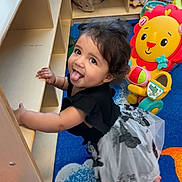 Zamira is registered to the contest to win money with this photo: child, toddler, playroom, toy, colorful, carpet, smiling, tongue_out, wooden_shelf, stuffed_animal, lion_toy, black_clothing, white_skirt, curly_hair, indoor, playful, happy, person, floor, furniture