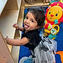 child, toddler, playroom, toy, colorful, carpet, smiling, tongue_out, wooden_shelf, stuffed_animal, lion_toy, black_clothing, white_skirt, curly_hair, indoor, playful, happy, person, floor, furniture