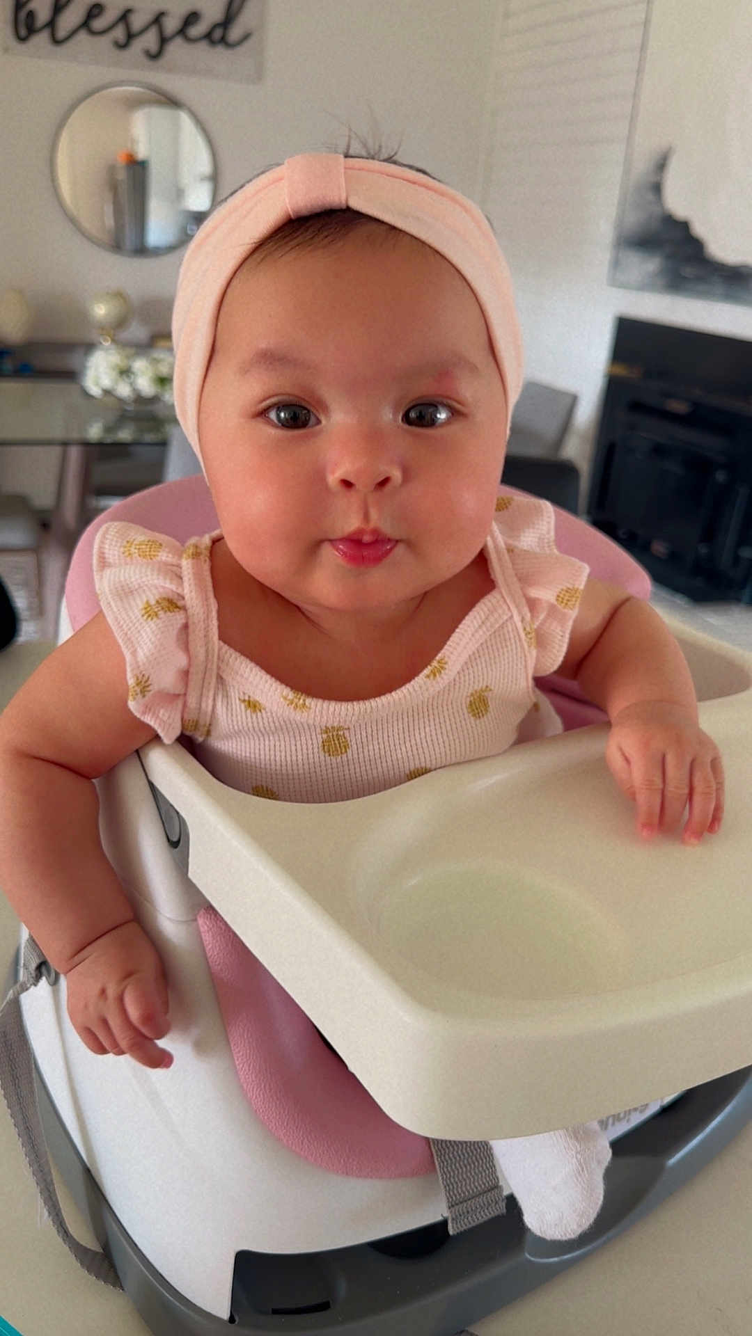 Yarely is registered to the contest to win money with this photo: baby, infant, high_chair, headband, pink_clothing, tongue_out, cute, big_eyes, chubby_cheeks, smiling, hands, face, eyes, dining_room, mirror, decor, wall_art, home_interior, tray, seat