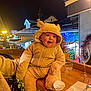 baby, building, christmas_decor, cozy, festive_lights, hooded_suit, infant, lamp_post, nighttime, person_arm, plush_outfit, portrait, railing, restaurant_balcony, smile, surprised_expression, warm_lighting, water_glass, wine_glass, wooden_table